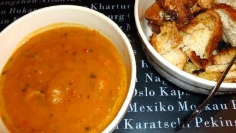 Eine cremige Kürbis-Kartoffelsuppe in einer Schüssel steht neben einer Portion gerösteter Brötchenwürfel auf einem Tisch.