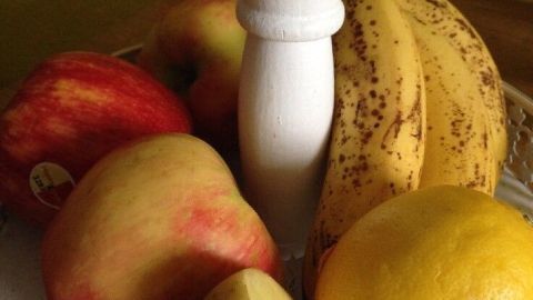 Ein Korb mit Obst, darunter eine rohe Kartoffel, frische Äpfel, eine Banane und eine Zitrone, stehen im Raum.