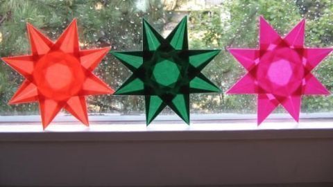 Bunte, transparente Sterne aus Buchfolie hängen am Fenster und strahlen durch das Tageslicht, das durch die Blätter scheint.
