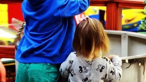 Zwei Kinder stehen an einem Spielplatz, während das eine Kind einen Eimer hebt und das andere zuschaut.
