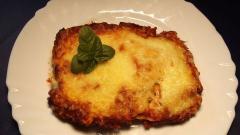 Ein Stück Lasagne mit geschmolzenem Käse liegt auf einem weißen Teller, dekoriert mit einem frischen Basilikumblatt.