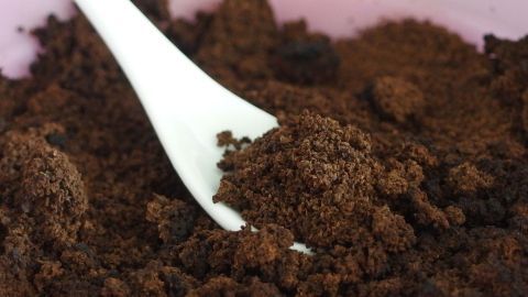 Ein weißer Löffel greift in ein Schälchen mit dunklem Kaffeepeeling, das auf einem rosa Untergrund liegt.