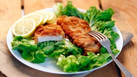 Schnitzel mit gewölbter Panier liegt auf einem Teller mit Salat und Zitronenscheiben, umgeben von einer Holzoberfläche.