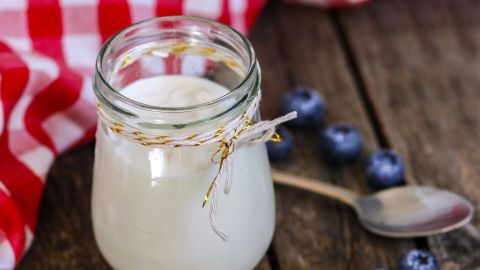 Ein Glas Naturjoghurt steht auf einem Holztisch, umgeben von frischen Blaubeeren und einem karierten Tuch.