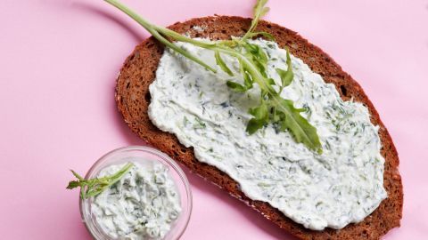 Auf einem Stück Vollkornbrot liegt ein Rucola-Dip, daneben ein Glas mit ähnlichem Dip, auf rosa Hintergrund.