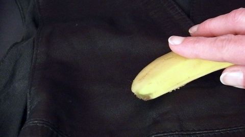 Eine Hand reibt eine Bananenschale über eine dunkle Lederoberfläche zur Pflege, im Hintergrund ist eine weitere Ledertextur sichtbar.