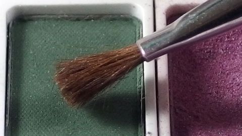 Eine Pinselspitze berührt grünen Lidschatten, während der lila Farbton nebenan im Makeup-Container liegt.