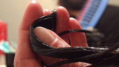 Eine Hand hält glänzende Haare, die mit Kokosöl behandelt sind, während im Hintergrund ein Laptop sichtbar ist.