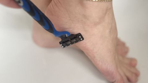 Eine Hand rasiert mit einem Rasierer einen Fuß, während der goldene Armreif in einem sauberen Badezimmer sichtbar ist.