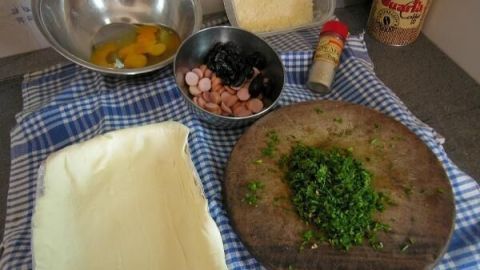 Auf einem Küchentisch sind Zutaten für einen Ei-Auflauf im Schlafrock vorbereitet: Eier, Schinken, Käse und frische Kräuter.