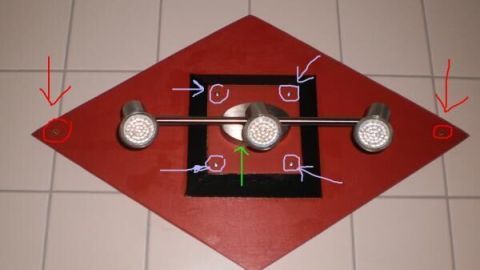 Eine Badlampe mit drei Strahlern ist mittig an einer rot-schwarzen rautenförmigen Platte auf Fliesen befestigt.