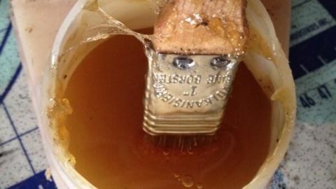 Ein Pinsel taucht in eine transparente Schale mit einer goldbraunen Gummilösung. Der Hintergrund ist eine Arbeitsfläche.