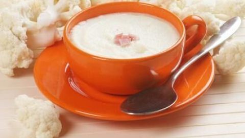 Blumenkohlsuppe in einer orangefarbenen Tasse auf einem passenden Teller mit Löffel, umgeben von Blumenkohlröschen.
