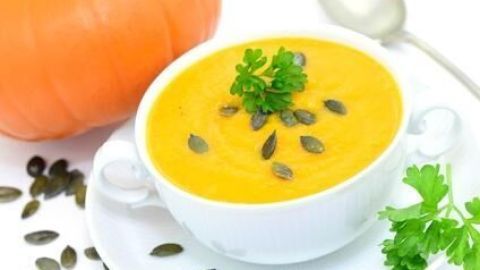 Eine Schale Kürbissuppe ist mit Kürbiskernen und Petersilie garniert, daneben liegt ein Löffel; im Hintergrund ein Kürbis.