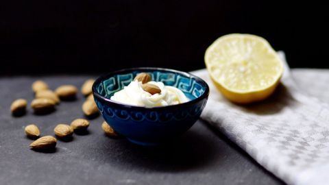 Eine kleine Schüssel mit Mandel-Mayonnaise steht auf einem dunklen Untergrund, umgeben von Mandeln und einer halbierten Zitrone.