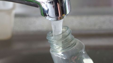 Eine Wasserleitung füllt eine Glasflasche, um Glasreiniger zu verdünnen. Im Hintergrund ist eine Arbeitsfläche sichtbar.