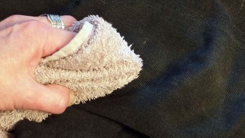 Eine Hand hält ein Tuch, das auf schwarzem Stoff reibt, um Waschmittelrückstände zu entfernen.