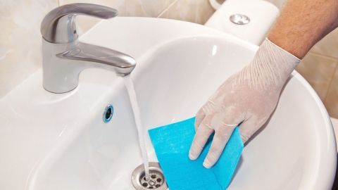 Eine Hand mit einem weißen Handschuh reinigt ein Waschbecken mit einem blauen Schwamm, während Wasser läuft.