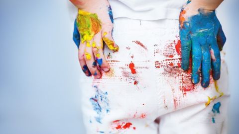 Bunte Hände mit blauen und gelben Farben berühren einen fleckigen weißen Overall im kreativen Arbeitsumfeld.