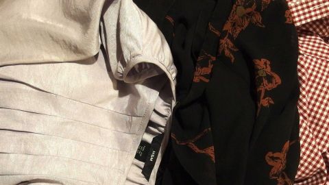 Kleidungsstücke liegen ungeordnet aufeinander. Ein graues Kleid von H&M, ein schwarzes, blumiges Kleidungsstück und ein rot-weiß kariertes Hemd.