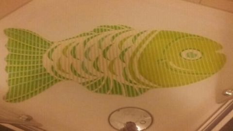 Eine grüne Badematte zeigt ein Fischdesign und liegt auf einem hellen Boden umgeben von Badezimmerfliesen.