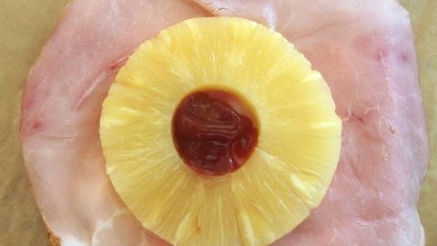 Auf einer Scheibe Schinken liegt eine Ananasscheibe mit Ketchup statt einer Kirsche, umgeben von einer neutralen Fläche.
