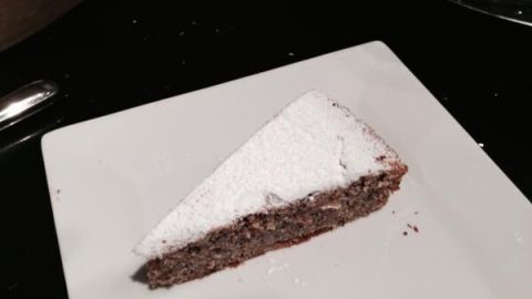 Ein Stück Haselnusskuchen liegt auf einem weißen Teller, bestäubt mit Puderzucker, auf einer dunklen Tischoberfläche.