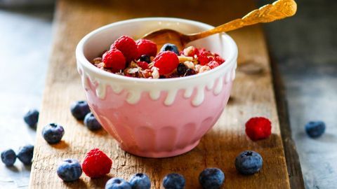 Eine pinke Schüssel voller Knuspermüsli mit frischen Himbeeren und Heidelbeeren steht auf einem Holzbrett.