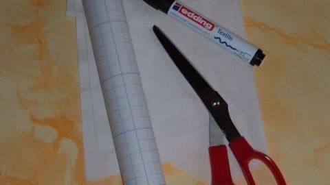 Ein gerolltes Blatt Papier steht neben einer Schere, einem Marker und einem weiteren Stück Stoff auf einem hellen Untergrund.