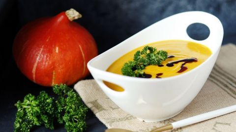 Eine weiße Schale mit Kürbis-Gemüse-Suppe steht auf einem Tisch, umgeben von einem Kürbis und frischem Grün.
