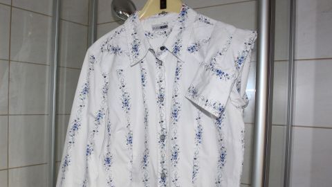 Mit diesem einfachen Trick kannst du Haarfärbemittelflecken auf Kleidung entfernen: Haarspray großzügig auf den Fleck sprühen, kurz einwirken lassen und dann waschen. Deine Bluse bleibt fleckenfrei und sauber! Ein weißes, geblümtes Hemd hängt an einem Bügel im Bad, umgeben von gefliesten Wänden und einer Duschkabine.