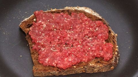 Ein Bissen „Bemme“ mit rohem Gehacktes liegt in einer Pfanne, bereit zum Braten, auf einer dunklen Brotscheibe.