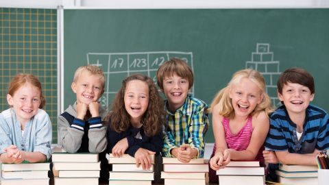 Kinder lachen hinter gestapelten Schulbüchern in einem Klassenzimmer, vor einer Tafel mit Matheschriftzügen.
