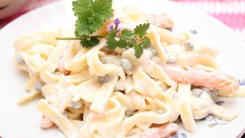 Ein Teller Nudelsalat liegt im Vordergrund, umgeben von buntem Hintergrund; er ist mit Kräutern dekoriert.