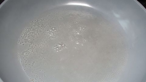Eine weiße Pfanne zeigt kleine Blasen, während Fleisch ohne Fett brät. Die Wärmequelle ist kaum sichtbar im Hintergrund.