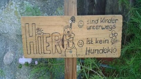Ein Holzschild steht in einer grünen Umgebung. Es weist darauf hin, dass hier Kinder unterwegs sind und kein Hundekot hinterlassen werden sollte.