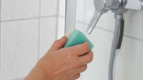 Eine Hand mit einem Schwamm entfernt Kalk von einer glatten Glasduschtür in einem Badezimmer.