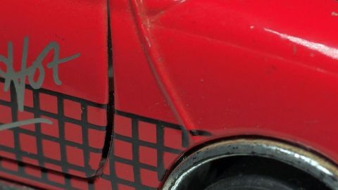 Ein rotes Auto mit einem Graffiti-ähnlichen Muster zeigt eine beschädigte Tür. Teerflecken sind sichtbar.