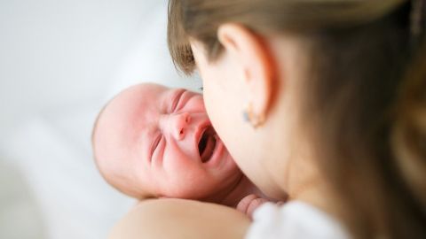 Ein weinendes Baby wird von einer Mutter beruhigt, während sie ihm liebevoll ins Gesicht schaut, in einem neutralen Raum.