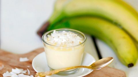 Ein Becher mit Kokos-Ananas-Quark steht auf einem Holzbrett, umgeben von Bananen, in einer hellen Küchenumgebung.