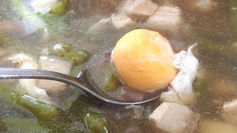 Eine Suppenkelle hält ein weich gekochtes Ei über einer klaren Suppe mit Gemüse und Fleischstücken, die dampft.