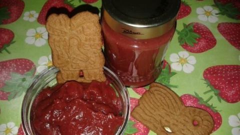 Eine Schüssel mit Erdbeerkonfitüre steht neben einem Glas, umgeben von Lebkuchen auf einem festlichen Untergrund mit Erdbeermuster.