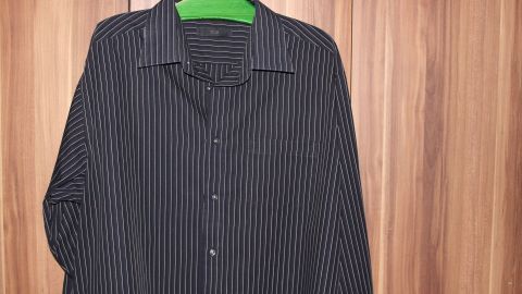 Mit einem einfachen Trick bleiben deine Hemden auf Reisen knitterfrei: Rolle statt falte sie und lege sie in den Koffer. So behält auch dieses gestreifte Hemd seinen frischen Look. Ideal für Geschäftsreisen! Ein gestreiftes, dunkles Hemd hängt glatt an einem grünen Bügel vor einem Holzwandschrank.