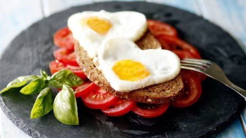 Ein Herzfrom-Spiegeleiergericht liegt auf Vollkornbrot, umgeben von frischen Tomatenscheiben und Basilikum.