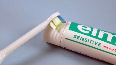 Eine Zahnbürste drückt auf eine Tube Zahncreme. Auf der Tube steht "elmex SENSITIVE mit Aminfluorid".