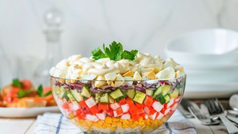 Ein farbenfroher Schichtsalat mit Gemüse und Eiern in einer Glasform steht auf einem Tisch, umgeben von Tellern und Besteck.