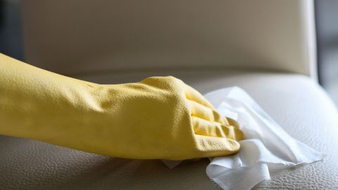 Eine Hand in einem gelben Handschuh wischt mit einem Tuch einen weißen Kunstlederstuhl in einem hellen Raum sauber.