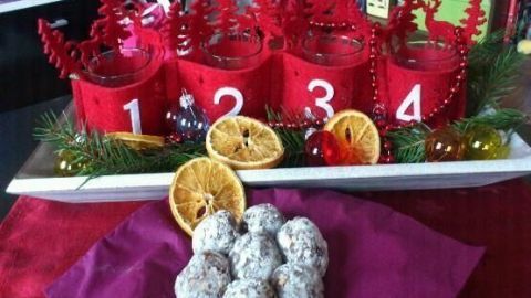 Kleine Kuchen-Kugeln liegen auf lila Stoff vor einem Adventskranz mit roten Kerzenhaltern (1-4), Tannenzweigen und Orangenscheiben.