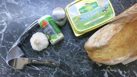 Entdecke, wie einfach es ist, deine eigene Knoblauchbutter herzustellen! Alles, was du brauchst, ist frischer Knoblauch, Butter, Kräuter und etwas Brot. Perfekt zum Dippen und Genießen – so machst du deine Mahlzeiten noch aromatischer. Knoblauch und Butter neben einer Knoblauchpresse, Gewürzstreuer und Brot auf einer schwarzen Marmor-Tischplatte. Text: "Kerrygold extri."