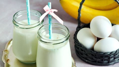 Zwei Gläser mit Protein-Shake aus Magerquark stehen auf einem Tablett, umgeben von Bananen und Eiern im Hintergrund.